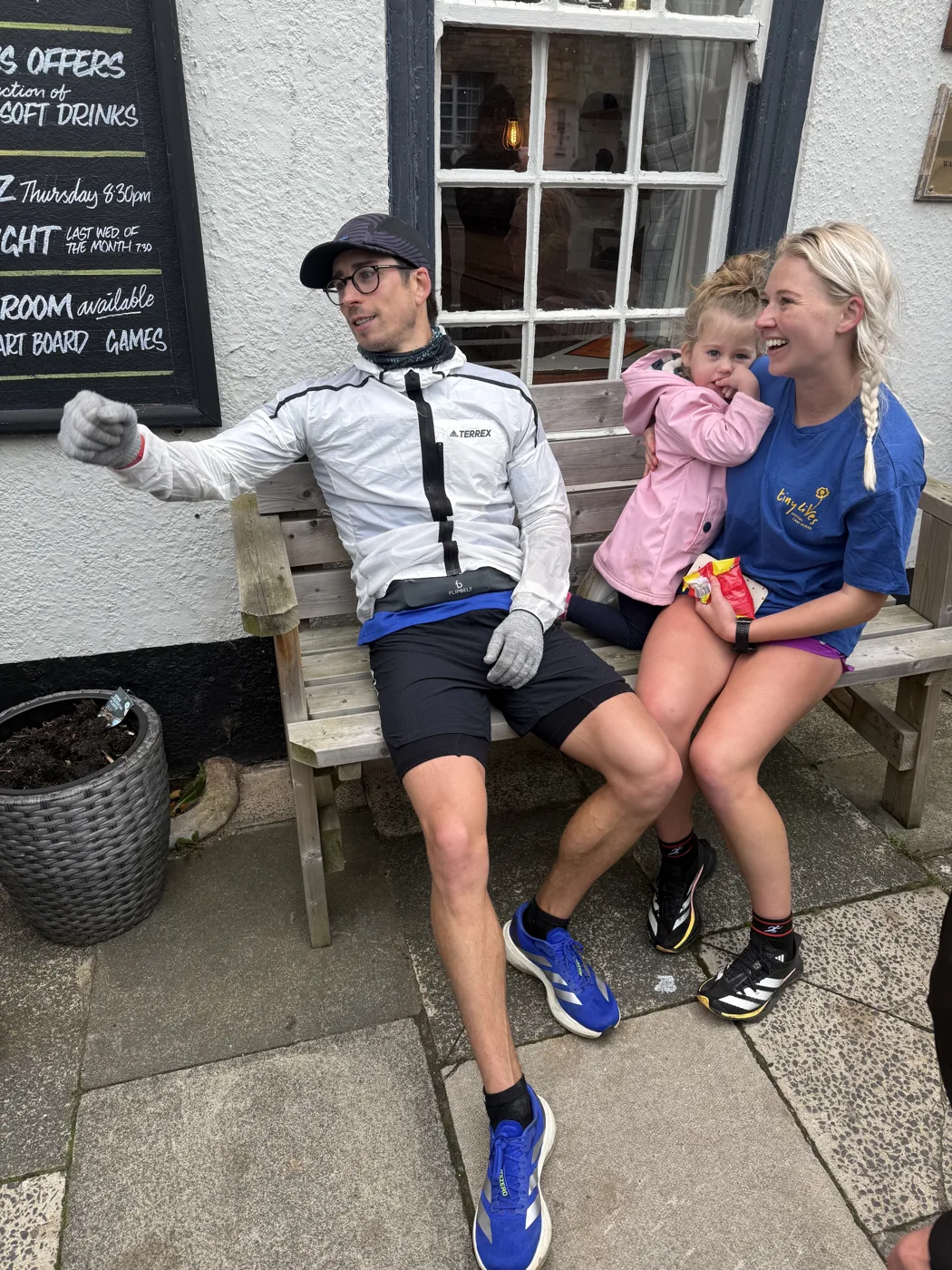 A runner in a cap, gloves and a white running jacket resting on a wooden bench outside a pub, beside a woman in a blue Tiny Lives T-shirt holding a small child in a pink coat